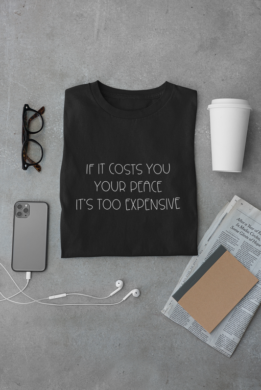 If It Costs You Your Peace It's Too Expensive T-Shirt in black with dainty white bold text. This is the front view of the shirt folded on a gray cement background with a pair of glasses, phone and headphones, newspaper, journal and white coffee cup surrounding it.