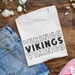 Minnesota Vikings - T-Shirt in white with black writing. Shirt is folded on a wooden background next to a pair of jeans, pink tennis shoes, headphones and white flowers.