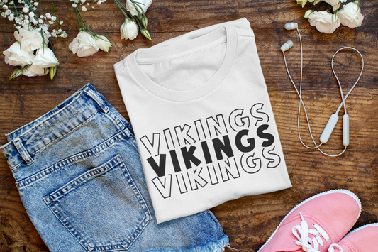 Minnesota Vikings - T-Shirt in white with black writing. Shirt is folded on a wooden background next to a pair of jeans, pink tennis shoes, headphones and white flowers.