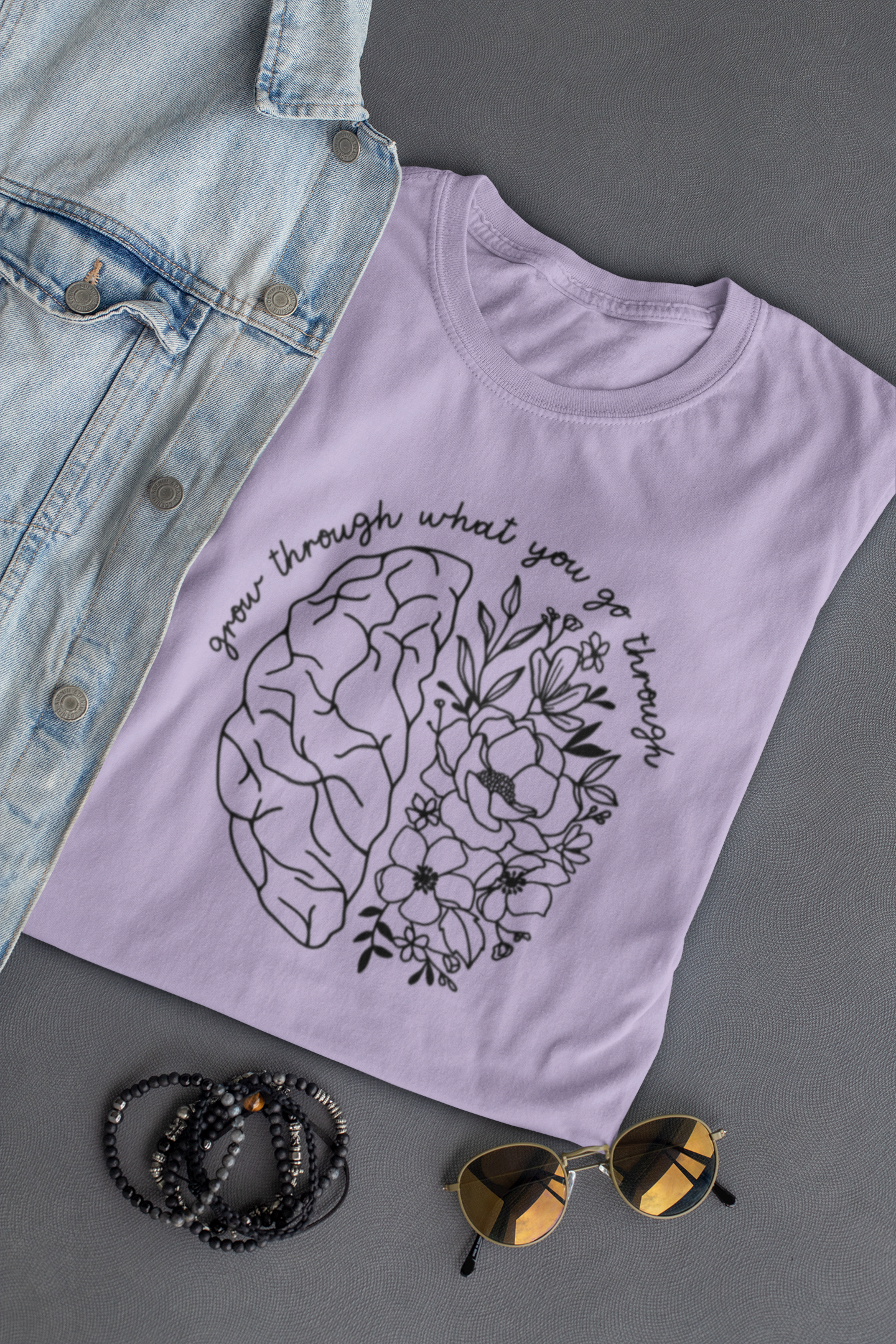 Grow Through Mental Health Brain T-Shirt in purple with a black design on the front. The design has a brain with the left side looking like a brain and the right side being flowers. At the top of the design has a cursive font that says "grow through what you go through". This is the front view of the shirt on a gray background, folded with a jean jacket, a pair of sunglasses and bracelets next to it.