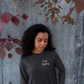 Not Everything That Weighs You Down - Pull Over Sweatshirt in charcoal gray. The back has a white cursive saying of "not everything that weighs you down is yours to carry" with the front corner pocket saying in a cursive font in white of "keep smiling". This is the front view of the sweatshirt on a model up against a wooden wall with leaves above her.