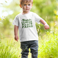 Cutest Clover In The Patch T-Shirt for youth and toddler in white. Adorable saying in green with twinkles and 4 leaf clovers. This is the front view of the shirt on a little boy looking at the camera, walking through a green patch.