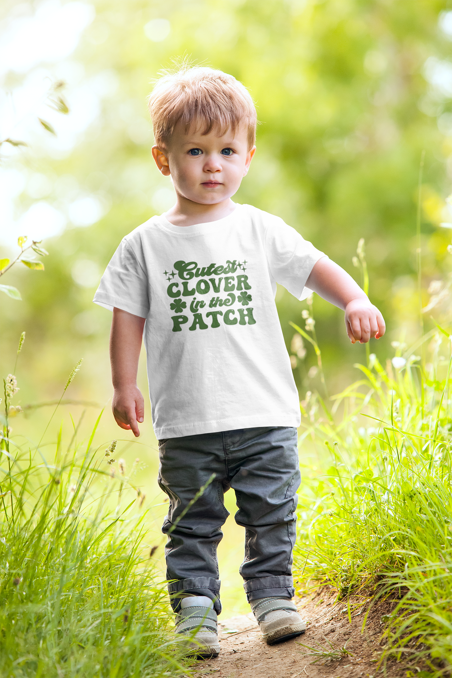 Cutest Clover In The Patch T-Shirt for youth and toddler in white. Adorable saying in green with twinkles and 4 leaf clovers. This is the front view of the shirt on a little boy looking at the camera, walking through a green patch.