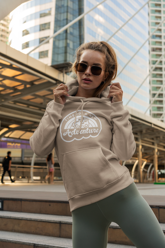 Time For A New Adventure Hooded Sweatshirt. Design is in a cloud bubble with time for a new adventure at the top curved with an off road vehicle in the middle with a rainbow behind it, and adventure below it. This the design in a white on the sandstone tan hoodie. A female model is wearing the sweatshirt with green leggings, sun glasses on and her hands holding the hood. She stands in the front a subway station in the middle of a city.