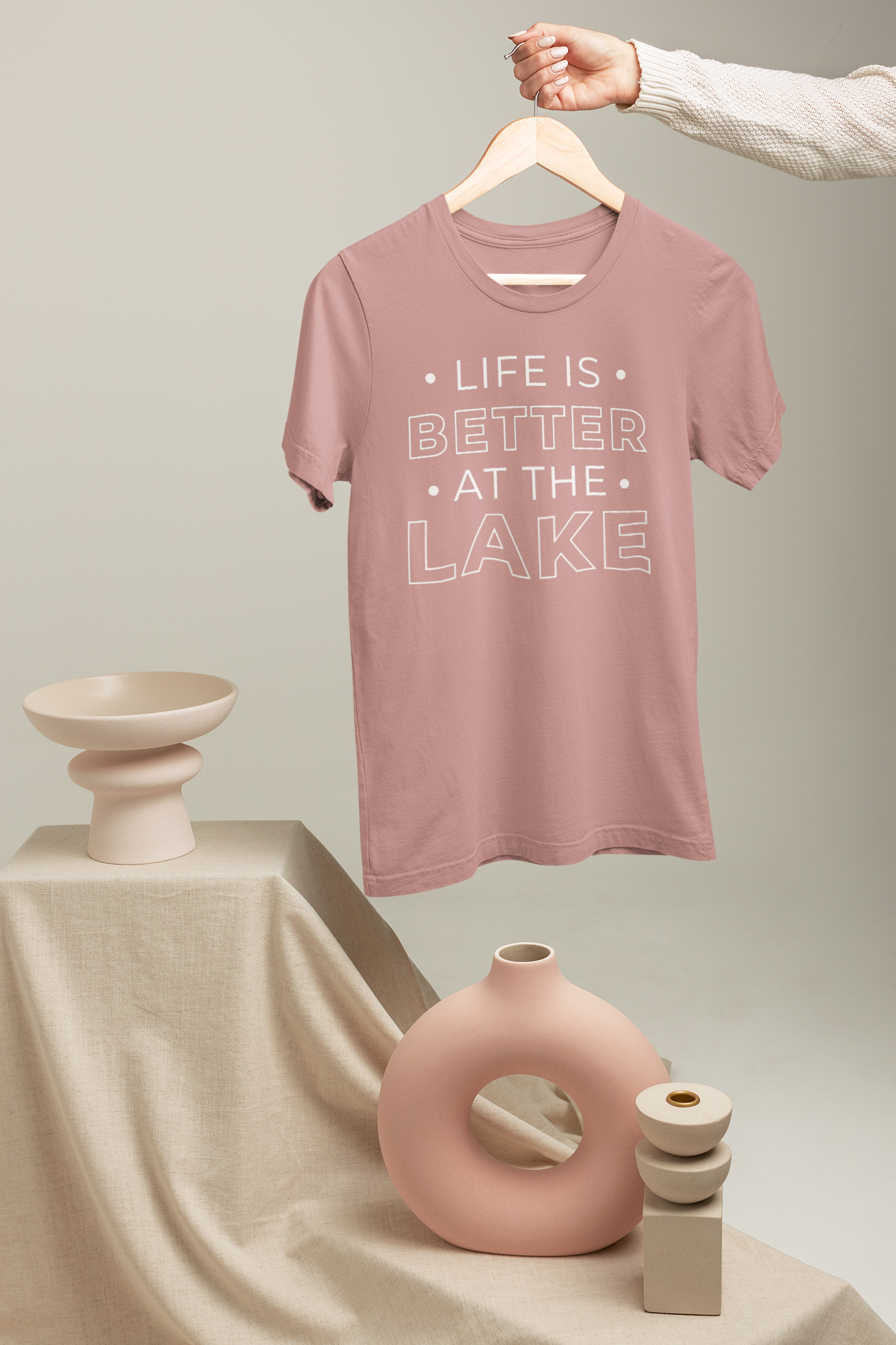 Life Is Better By The Lake - T-Shirt in Mauve with white writing hanging on a hanger and someone holding said hanger. Styled items below the item on a staircase like set.
