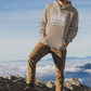 Time For A New Adventure Hooded Sweatshirt. Design is in a cloud bubble with time for a new adventure at the top curved with an off road vehicle in the middle with a rainbow behind it, and adventure below it. This the design in a white on the sandstone tan hoodie. The sweatshirt is on a man with a winter hat on, khakis and hiking boots on top of a mountain facing the sunlight.