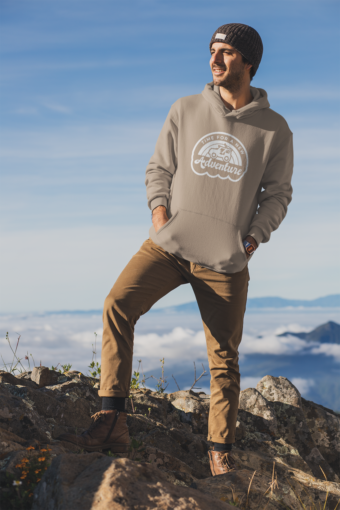 Time For A New Adventure Hooded Sweatshirt. Design is in a cloud bubble with time for a new adventure at the top curved with an off road vehicle in the middle with a rainbow behind it, and adventure below it. This the design in a white on the sandstone tan hoodie. The sweatshirt is on a man with a winter hat on, khakis and hiking boots on top of a mountain facing the sunlight.