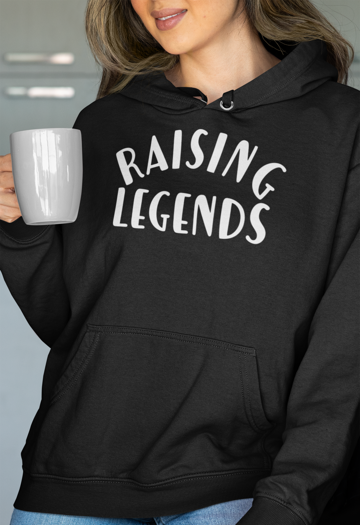 Raising Legends Black Sweatshirt Hoodie - Front ViewRaising Legends - Hooded Sweatshirt in black with white writing. Raising is curve at the top and legends is straight on the bottom. Features an innovative print process that raises the ink for a more defined design. This is the front view of the sweatshirt on a model holding a cup of coffee and posing.