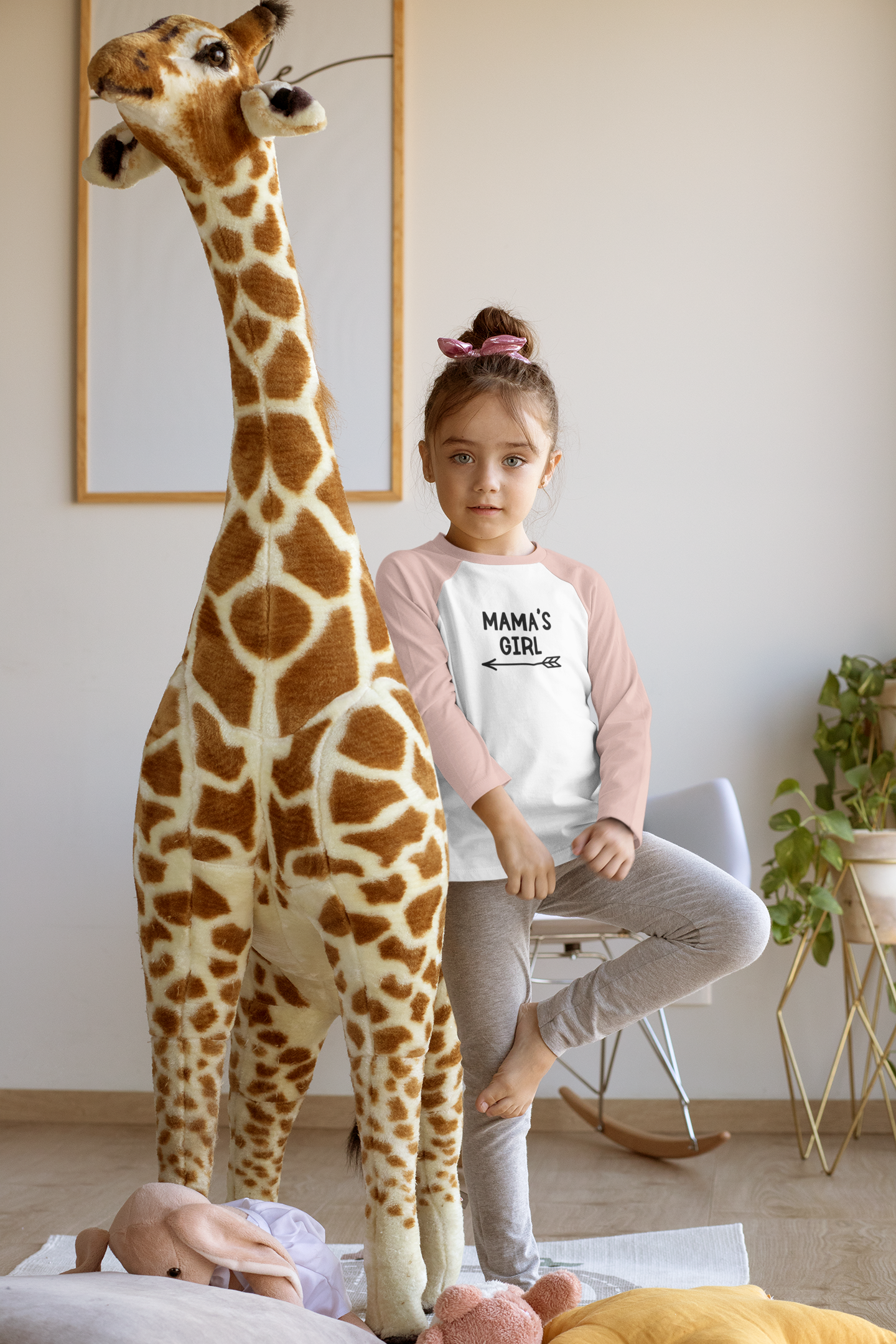 Mama's Girl - Three Quarter Sleeve T-Shirt. Sleeves are heather peach and base is white with the Mama's Girl writing in black with a cute arrow pointing to the left underneath. This is a picture of a young girl wearing the shirt in a playroom next to a giant giraffe, posing.