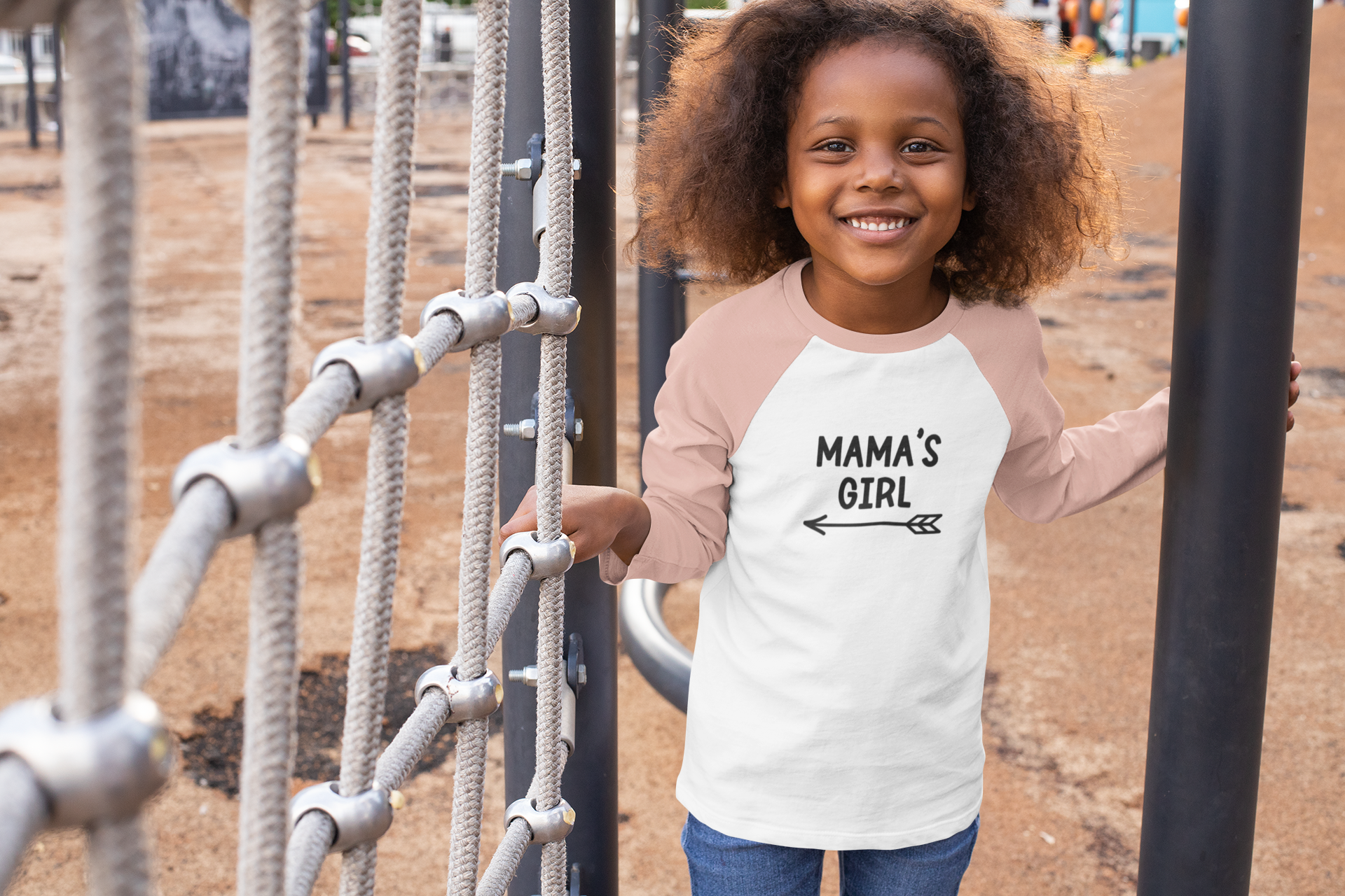 Mama's Girl - Three Quarter Sleeve T-Shirt. Sleeves are heather peach and base is white with the Mama's Girl writing in black with a cute arrow pointing to the left underneath. This is an image of a young girl wearing the shirt in a playground.
