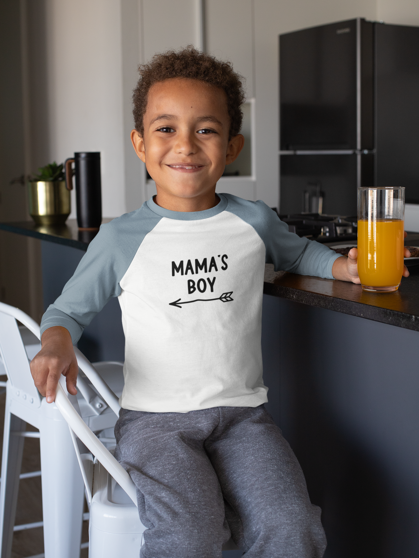 Mama's Boy - Three Quarter Baseball T-Shirt. Sleeves in a Heather light blue on the sleeves with a white base. Mama's Boy is in black with an arrow below it pointing to the left. This is the front view of the shirt on a little boy sitting at a kitchen counter drinking a glass of orange juice.