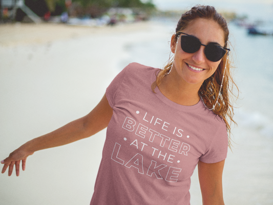 Life Is Better By The Lake - T-Shirt in Mauve with white writing  on a model at the lake with sunglasses on smiling