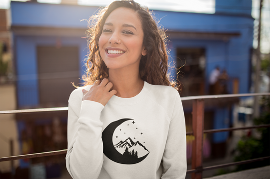 Moonlight Mountain - Pull Over Sweatshirt. The design is in black with a half moon, pine trees mountains and stars sitting inside the moon. This design is on a white sweatshirt. The image is of a girl with it on, sitting on at an outdoor patio smiling.