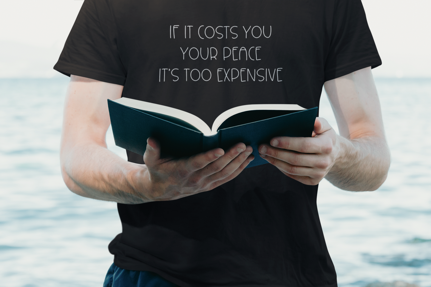 If It Costs You Your Peace It's Too Expensive T-Shirt in black with dainty white bold text. This is the front view of the shirt on a male model in front of water unclose with his hands out holding a book.