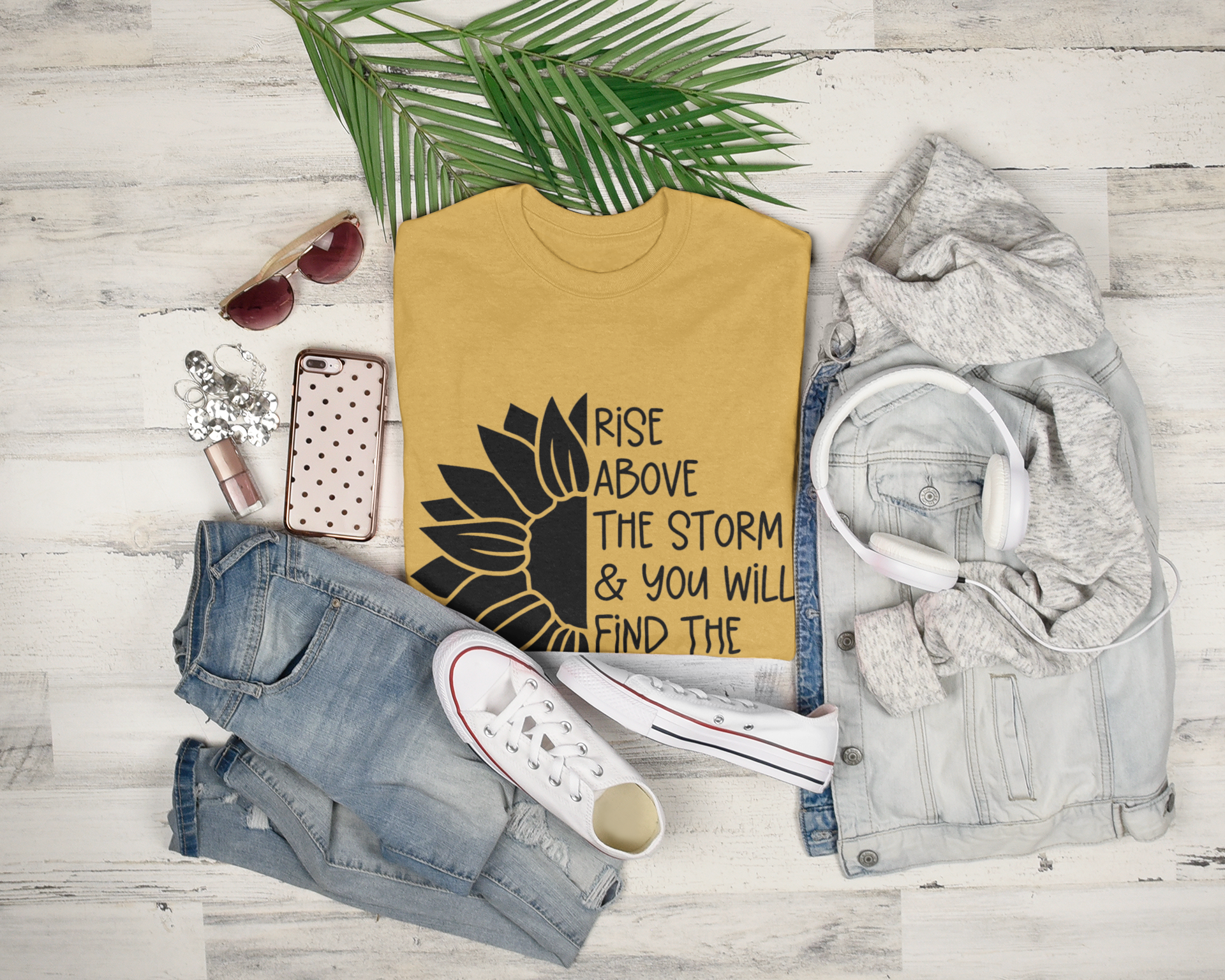 Rise Above The Storm Sunflower T-Shirt. The front has a half of a black sunflower on the left, with the right half having a quote of "rise above the storm and you will find the sunshine". This is a black design on a yellow shirt. This is the front view of the shirt folded on a wooden background stylized with other like items next to it.