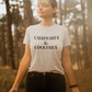 Campfires & Cocktails - T-Shirt in Heather oatmeal. The front of the shirt has a black bold font with campfires & cocktails on it. This is the front view of the shirt on a female model closing her eyes with her hands out beside her, standing in a forest area.