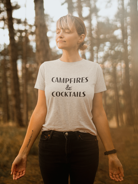 Campfires & Cocktails - T-Shirt in Heather oatmeal. The front of the shirt has a black bold font with campfires & cocktails on it. This is the front view of the shirt on a female model closing her eyes with her hands out beside her, standing in a forest area.