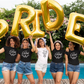 Floral Bride T-Shirt in white. The front has a bouquet of dainty flowers with the word "bride" written in the stems in a black design. This is the front view of the shirt on a bride with her girls standing in front of a pool holding up balloons writing bride. The girls next to her have black shirts on that so "I do crew"