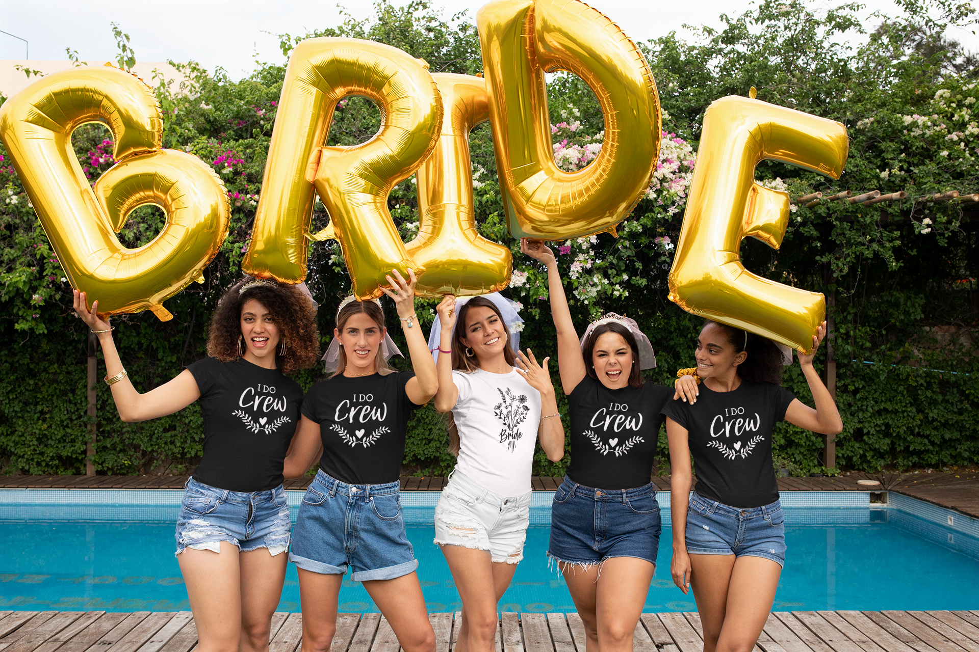 Floral Bride T-Shirt in white. The front has a bouquet of dainty flowers with the word "bride" written in the stems in a black design. This is the front view of the shirt on a bride with her girls standing in front of a pool holding up balloons writing bride. The girls next to her have black shirts on that so "I do crew"