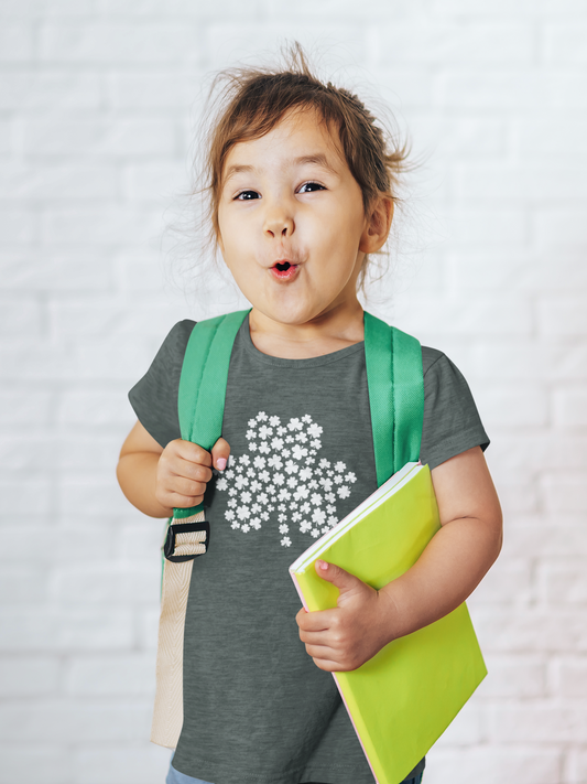Four-Leaf Clover T-Shirt in heather forest. The front of the t-shirt has a large four leaf clover with mini four leaf clovers making the shape of the larger clover in white. This is the front view of the shirt on a cute little girl making a cute face holding a backpack on her back and a green notebook in her left hand.