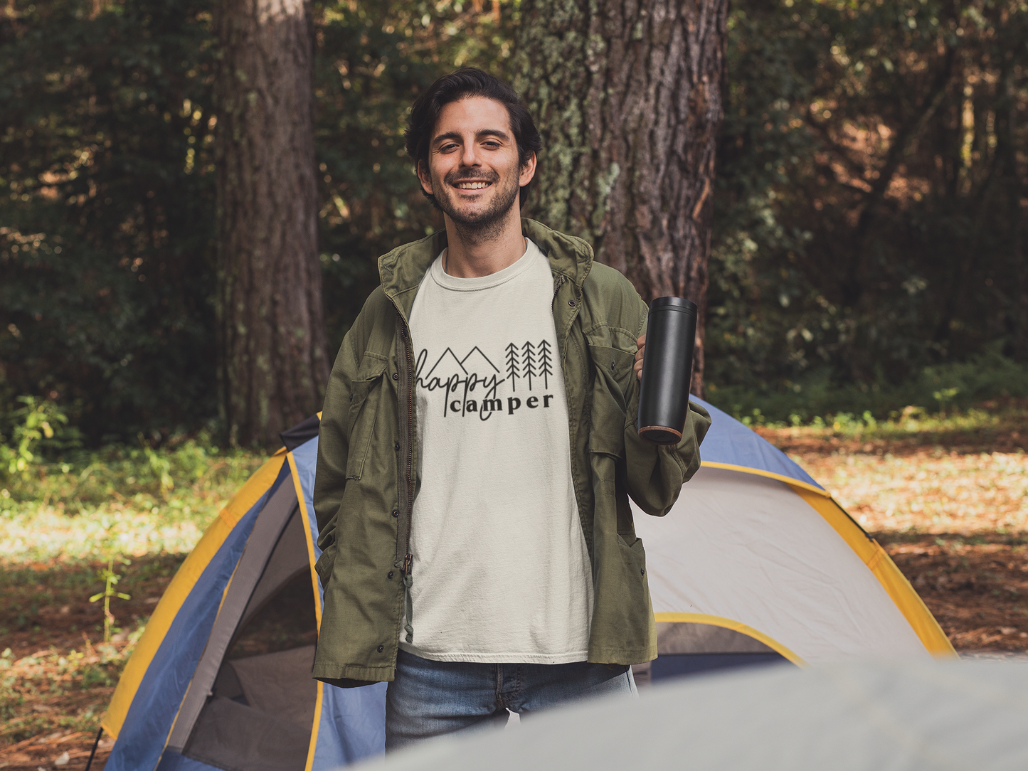 Happy Camper T-Shirt in Heather Oatmeal. The front has a cursive font "happy" with "camper" below it in a bold lowercase font. Two triangle mountains are above the word happy, and three pine trees are above the word camper. The design is in black. This is the front view of the shirt on a male model with a green jacket on holding up a mug and smiling in a woodsy scene with a camping tent behind him.