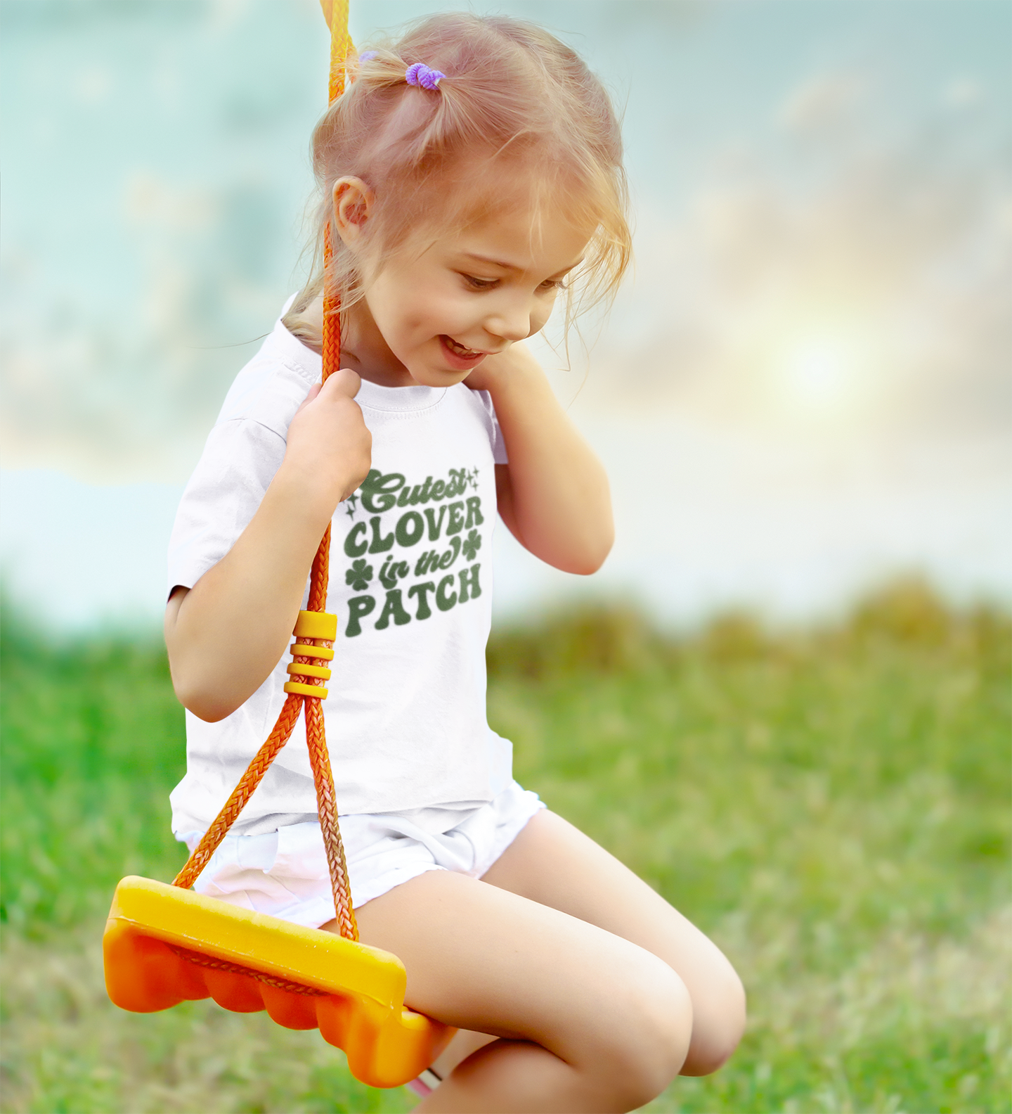 Cutest Clover In The Patch T-Shirt for youth and toddler in white. Adorable saying in green with twinkles and 4 leaf clovers. This is the front view of the shirt on a cute little girl swinging on a swing and smiling.