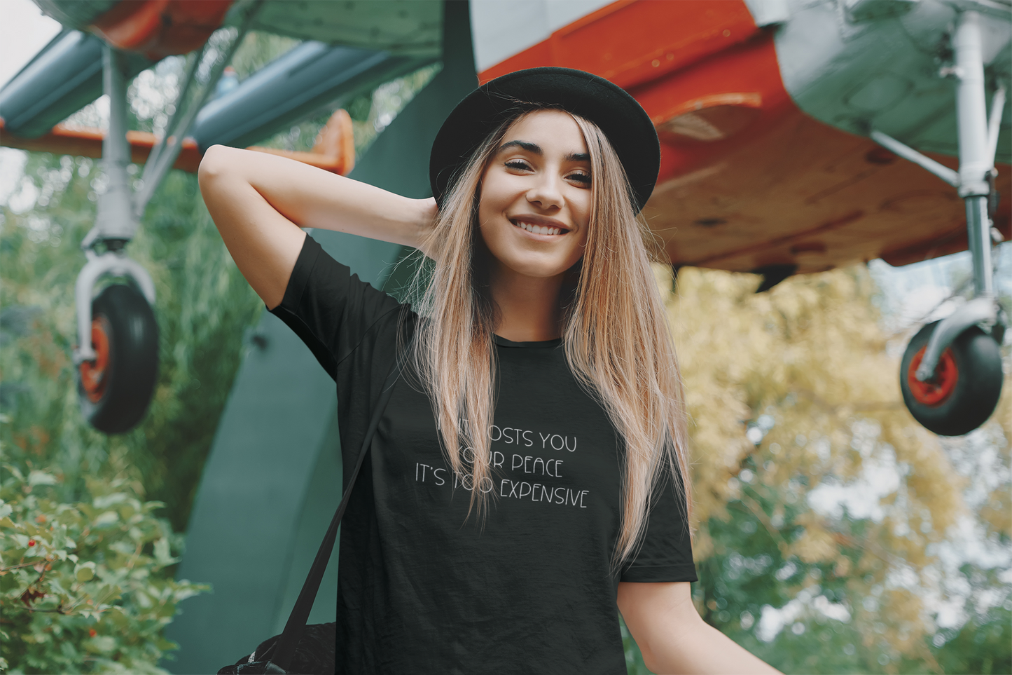 If It Costs You Your Peace It's Too Expensive T-Shirt in black with dainty white bold text. This is the front view of the shirt on a female model in a green area with a status airplane behind her. She is posing in front of it with a black fedora hat and a black bag hanging off her shoulder.