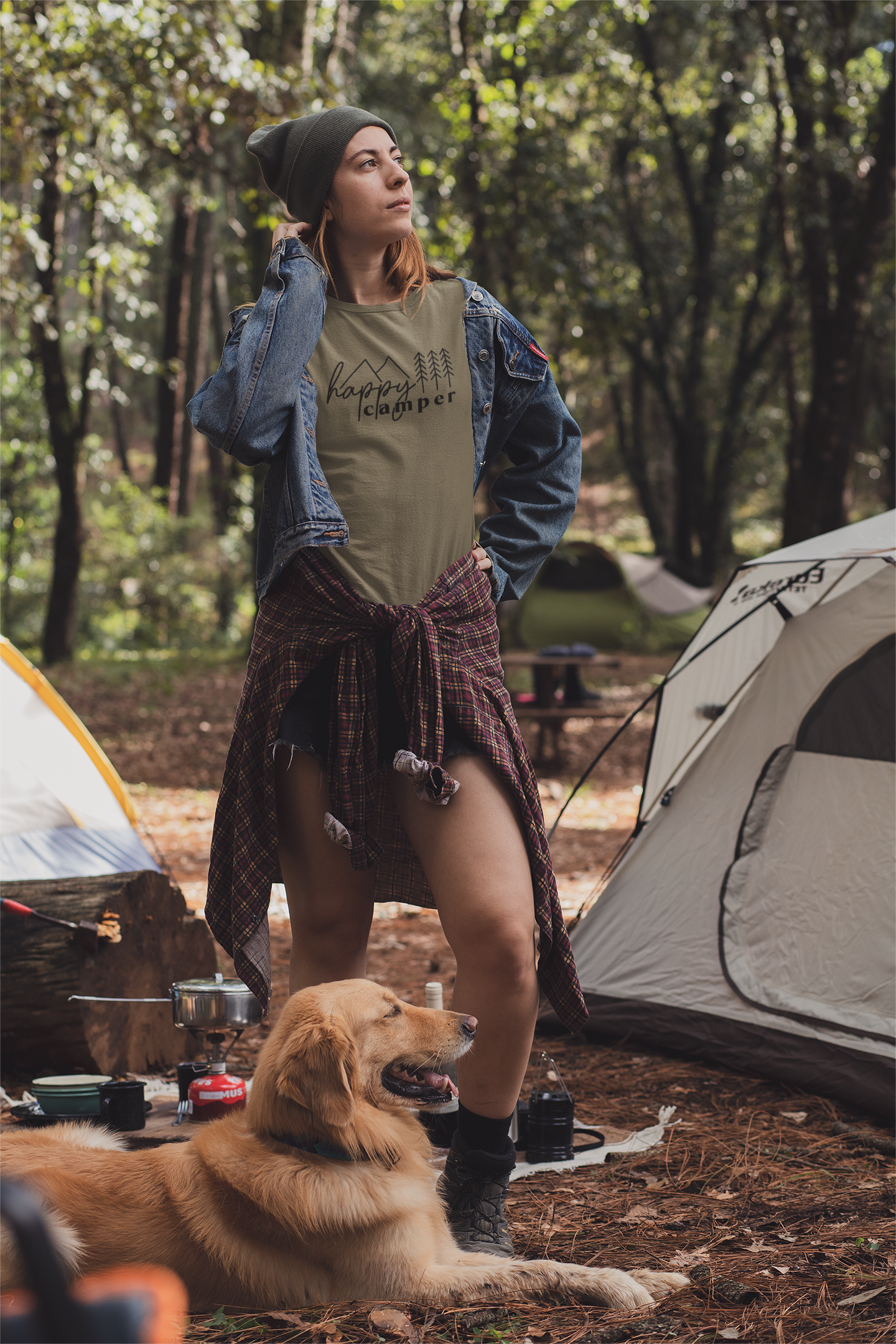 Happy Camper T-Shirt in Heather Olive. The front has a cursive font "happy" with "camper" below it in a bold lowercase font. Two triangle mountains are above the word happy, and three pine trees are above the word camper. The design is in black. This is the front view of the shirt on a female model posing looking up at the sky, with a dog in front of her legs in a woodsy area with tents beside her.