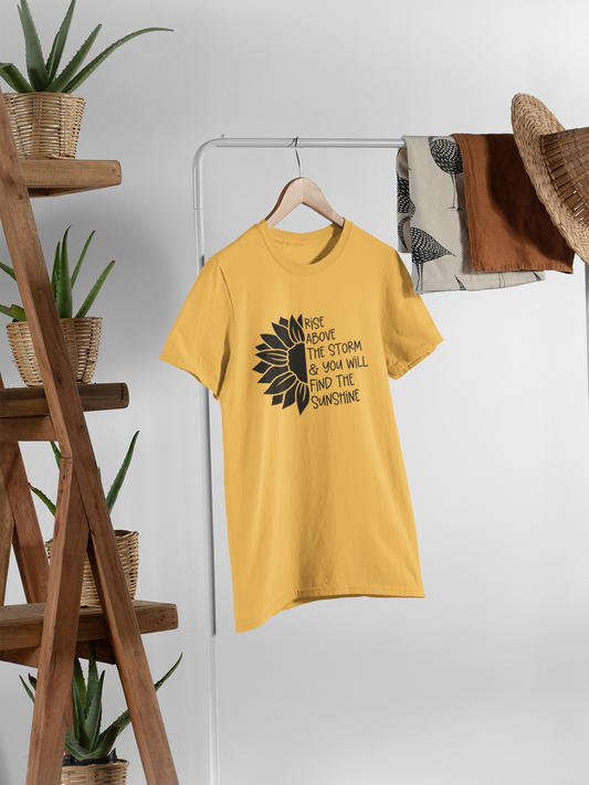 Rise Above The Storm Sunflower T-Shirt. The front has a half of a black sunflower on the left, with the right half having a quote of "rise above the storm and you will find the sunshine". This is a black design on a yellow shirt. This is the front view of the shirt hanging on a hanger on a clothing rack with items stylized around it.