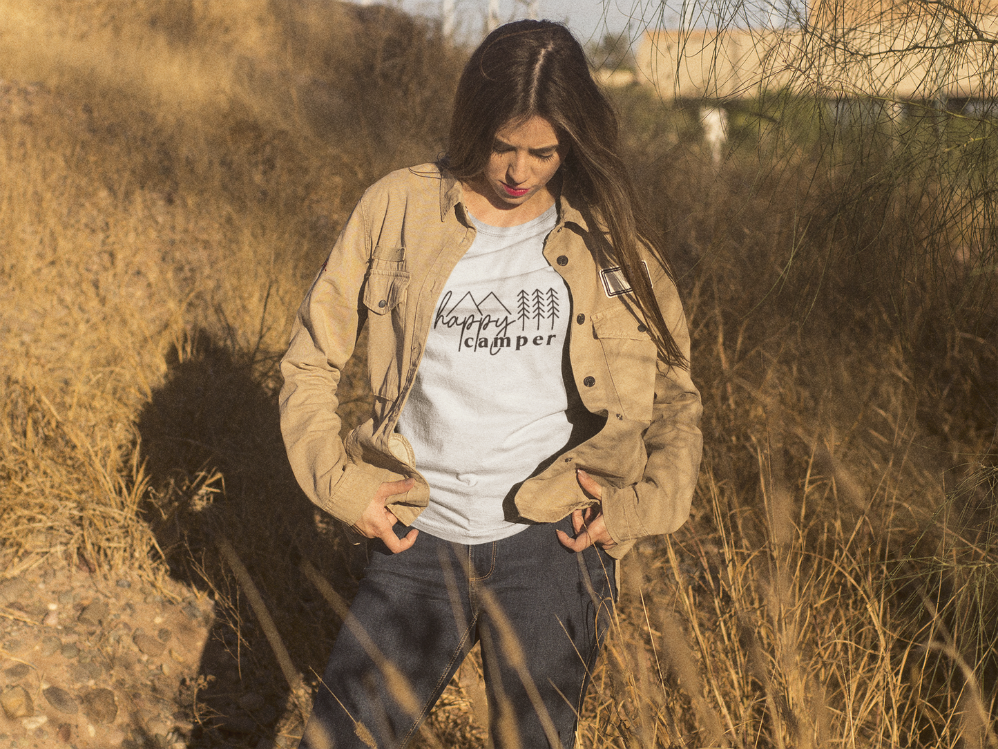 Happy Camper T-Shirt in Heather Oatmeal. The front has a cursive font "happy" with "camper" below it in a bold lowercase font. Two triangle mountains are above the word happy, and three pine trees are above the word camper. The design is in black. This is the front view of the shirt on a female model in the middle of a tan wooded field. 