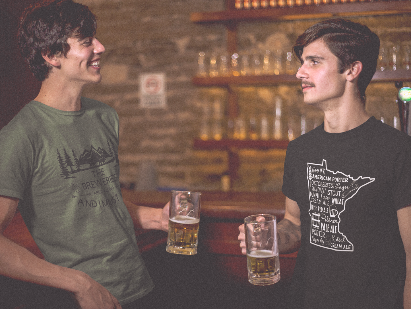 2 male models drinking beer in a bar laughing and having a good time. The left model is wearing a green t-shirt with a black design "the breweries are calling and I must go" the right hand model is wearing a Minnesota Beer Shirt with different kinds of beers inside the Minnesota design