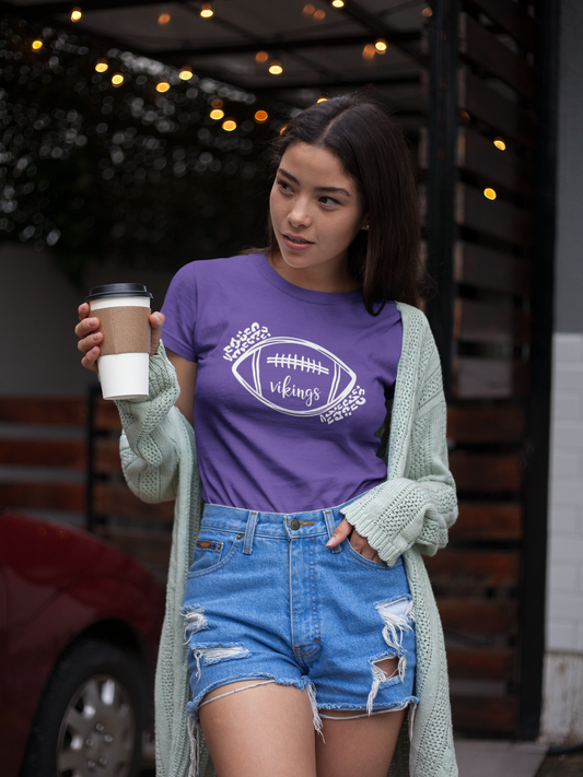 Leopard Vikings Graphic - Purple T-Shirt with white writing. The front features a sketchy outline of a football with "vikings" in a script font inside the bottom of the football with leopard print enclosing the top left and bottom right of the football. This is the front view of the t-shirt on a female model wearing jean shorts with the shirt tucked in, a green sweater with a cup of coffee in her hand.
