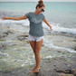 Beach Vibes T-Shirt on a female model oceanside walking through the water. Model has her hair in a bun, sunglasses on with the beach vibes shirt and light jean shorts.