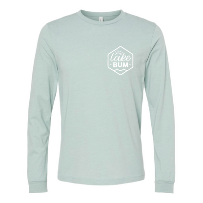 Life is Better at the Lake - Long Sleeve T-Shirt (front view) with patch on front that says Lake BumLife is Better at the Lake - Long Sleeve T-Shirt in heather dusty blue. The back of the shirt has a white design with the saying "Life is better at the lake" with lake waves, pine trees, and a loon all squared into a frame. The front has the saying "lake bum" with a lake wave and sun all boxed into a hexagon on the small square pocket (right). This is the front view of the shirt.