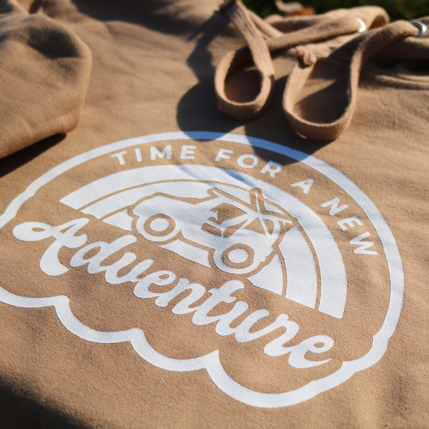 Time For A New Adventure Hooded Sweatshirt. Design is in a cloud bubble with time for a new adventure at the top curved with an off road vehicle in the middle with a rainbow behind it, and adventure below it. This the design in a white on the sandstone tan hoodie. This is a stylized up close shot of the design on the sweatshirt.