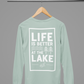 Life is Better at the Lake - Long Sleeve T-Shirt in heather dusty blue. The back of the shirt has a white design with the saying "Life is better at the lake" with lake waves, pine trees, and a loon all squared into a frame. The front has the saying "lake bum" with a lake wave and sun all boxed into a hexagon on the small square pocket (right). This is the back view of the shirt hanging on a natural wooden hanger hanging on a clothing rack.