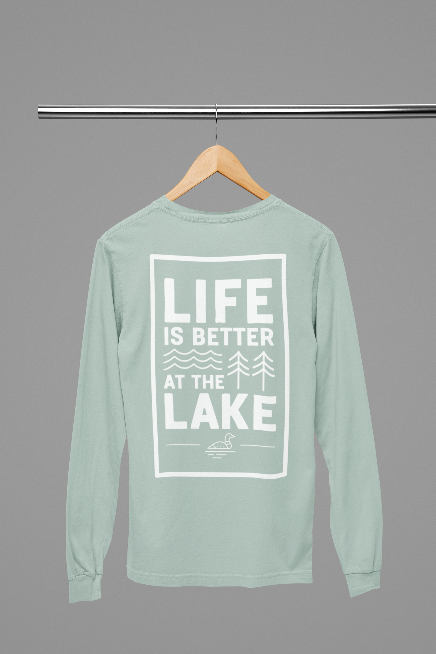 Life is Better at the Lake - Long Sleeve T-Shirt in heather dusty blue. The back of the shirt has a white design with the saying "Life is better at the lake" with lake waves, pine trees, and a loon all squared into a frame. The front has the saying "lake bum" with a lake wave and sun all boxed into a hexagon on the small square pocket (right). This is the back view of the shirt hanging on a natural wooden hanger hanging on a clothing rack.