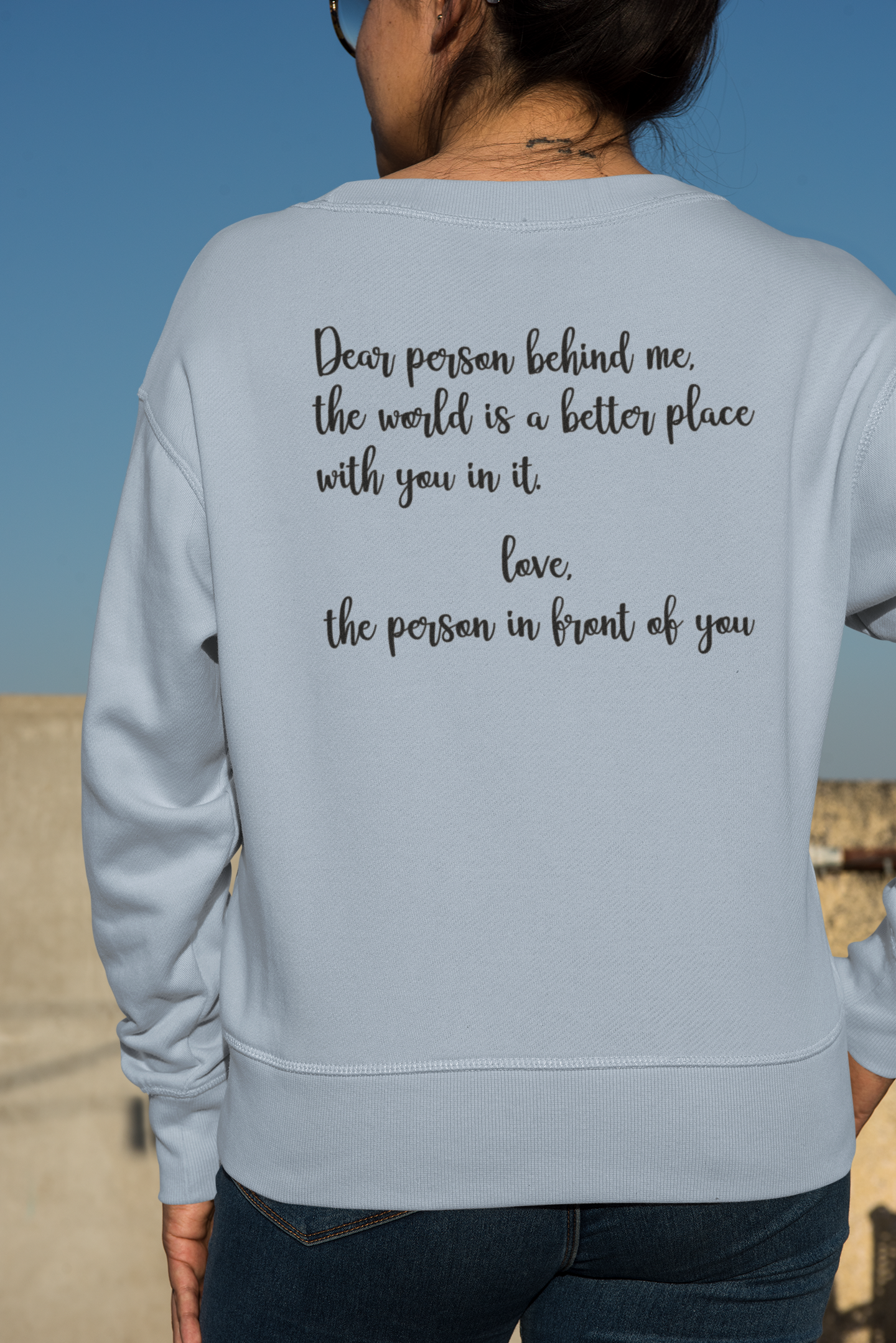 Dear Person Behind Me - Pull Over Sweatshirt in light blue. Designs are in black. The back says "Dear person behind me, the world is a better place with you in it. Love the person in front of you" in a cursive design. The front design is on the right hand side, as a small pocket square with the saying "be kind to your mind" in cursive with a heart and semi colon surrounding it. This is the back view of the sweatshirt on a model in an outdoor setting.