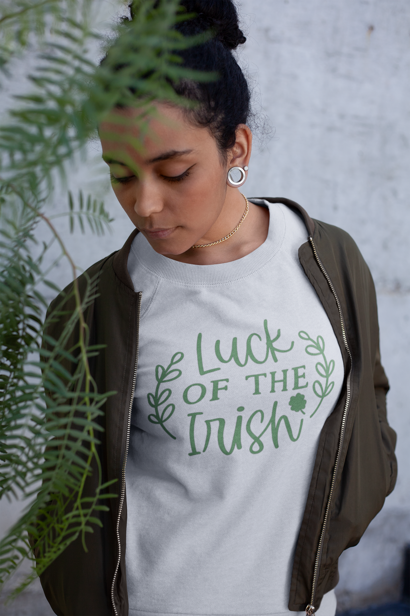 Luck Of The Irish - Pull Over Sweatshirt in a heather light gray. Luck Of The Irish quote on the sweatshirt is a medium green with cute leaves enclosing the design and a small four leaf clover. This is a stylized picture of a women wearing the sweatshirt with a green bomber jacket over the top of it, with some greenery in the front of her.