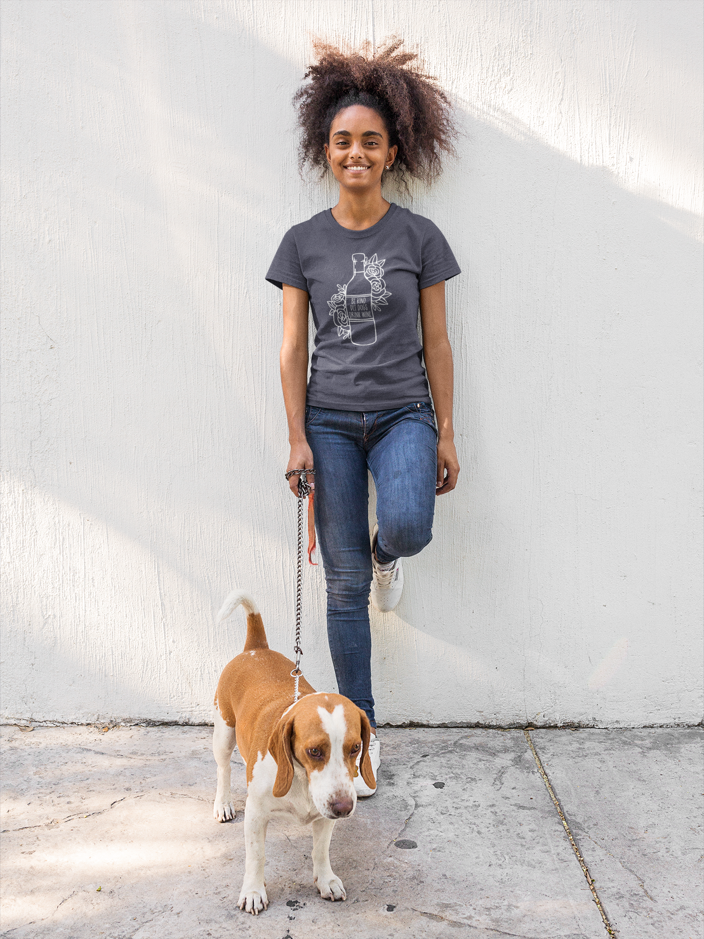 Be Kind, Pet Dogs, Drink Wine - T-Shirt on a navy shirt with white design. The design is an outline of a wine bottle with a variety of flowers and vines sketched beside the bottle, with the saying "be kind, pet dogs, drink wine" inside the wine label. This is the front view of the shirt on a model with her hair up standing against a white wall and side walk with a dog on a leash in her hand.