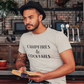 Campfires & Cocktails - T-Shirt in Heather oatmeal. The front of the shirt has a black bold font with campfires & cocktails on it. This is the front view of the shirt on a male model holding a book in a bar like setting with black lid hat and light dem jeans.
