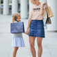 Mama - T-Shirt in heather prism peach. Mama is in a black design with an arrow below it pointing to the right. This is an image of a mom and her daughter shopping with her holding shopping bags. Mom has the Mama shirt on with denim jean skirt and tennis, with the little girl holding her hand wearing a light purple dress, gray sweater and black converse tennis shoes.