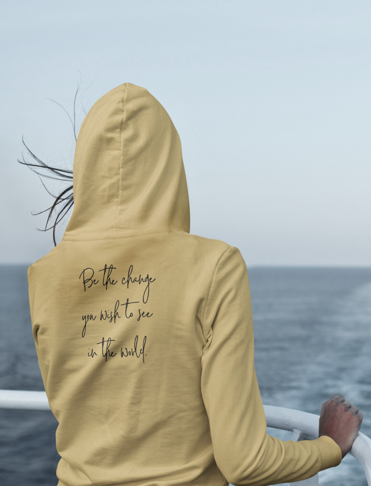 Be The Change Hooded Sweatshirt in a light yellow. The back has a black cursive writing saying "be the change you wish to see in the world." The front has a small flower with stem in black - pocket style. This is the back view of the sweatshirt on a model on the back of a boat with her hood up looking out at the water, and her hair waving in the wind.