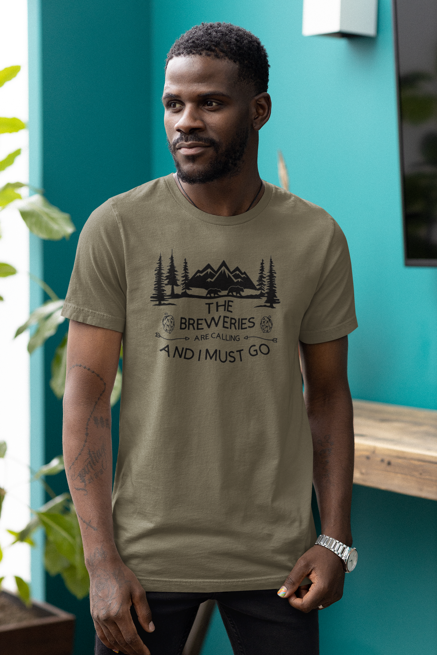 The Breweries Are Calling And I Must Go - T-Shirt in a heather Olive Garden. Black Design on the front with the saying "the breweries are calling and I must go" with arrows pointing at "are calling" hoops surrounding the "breweries" and pine trees, mountains and 2 black bears above the saying. This is the front view of the shirt on a male model hanging out in a brewery setting.