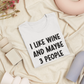 I Like Wine & Maybe 3 People - White T-Shirt in black writing. This is an image of the t-shirt folded in a square on a cream blanket, with a stringy pink flower and cream, blush pink, and light blue objects next to it.