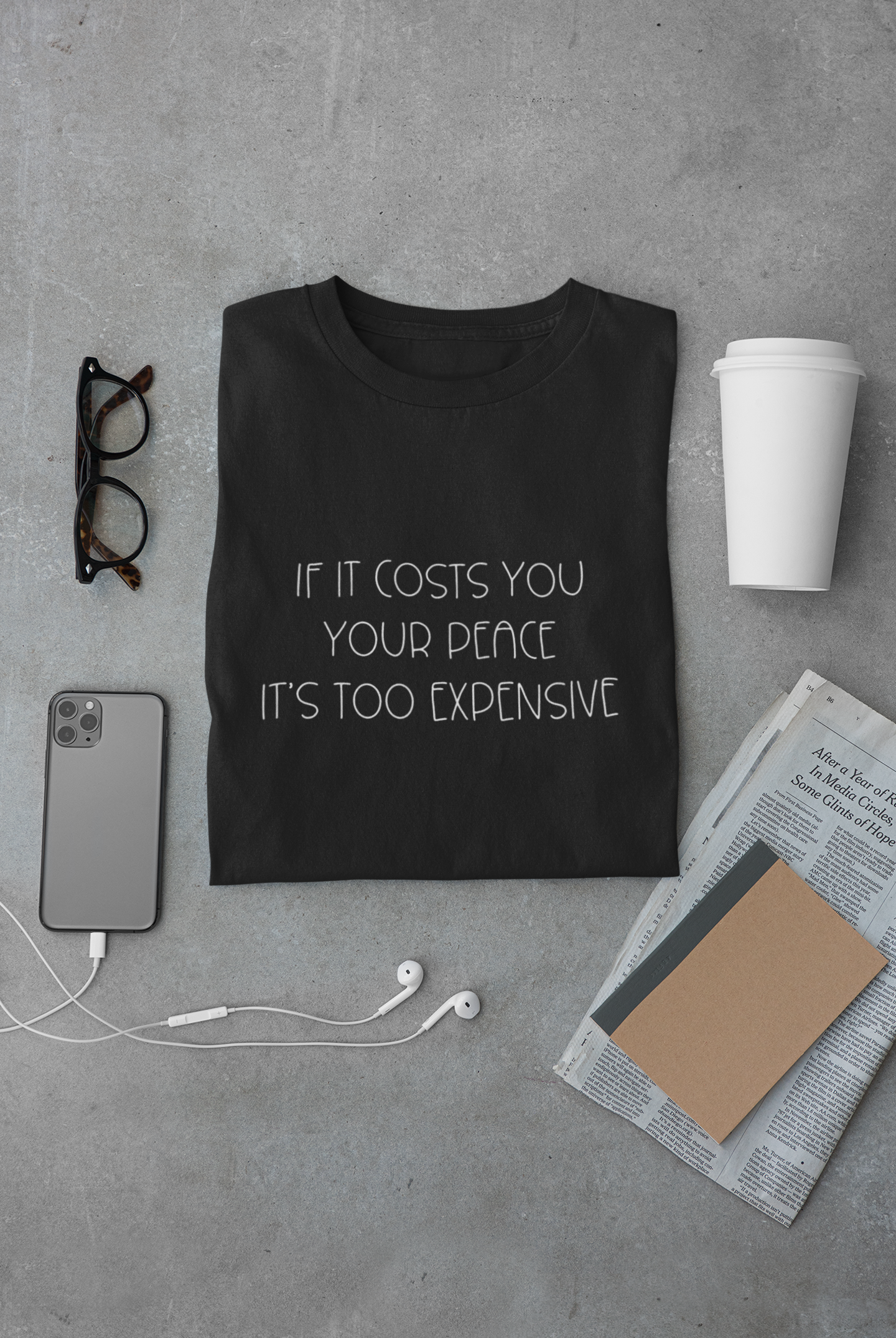 If It Costs You Your Peace It's Too Expensive T-Shirt in black with dainty white bold text. This is the front view of the shirt folded on a gray cement background with a pair of glasses, phone and headphones, newspaper, journal and white coffee cup surrounding it.