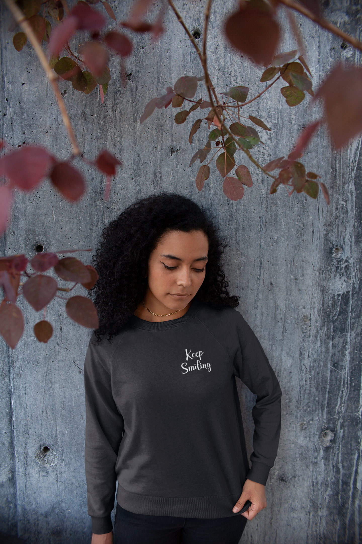 Not Everything That Weighs You Down - Pull Over Sweatshirt in charcoal gray. The back has a white cursive saying of "not everything that weighs you down is yours to carry" with the front corner pocket saying in a cursive font in white of "keep smiling". This is the front view of the sweatshirt on a model up against a wooden wall with leaves above her.