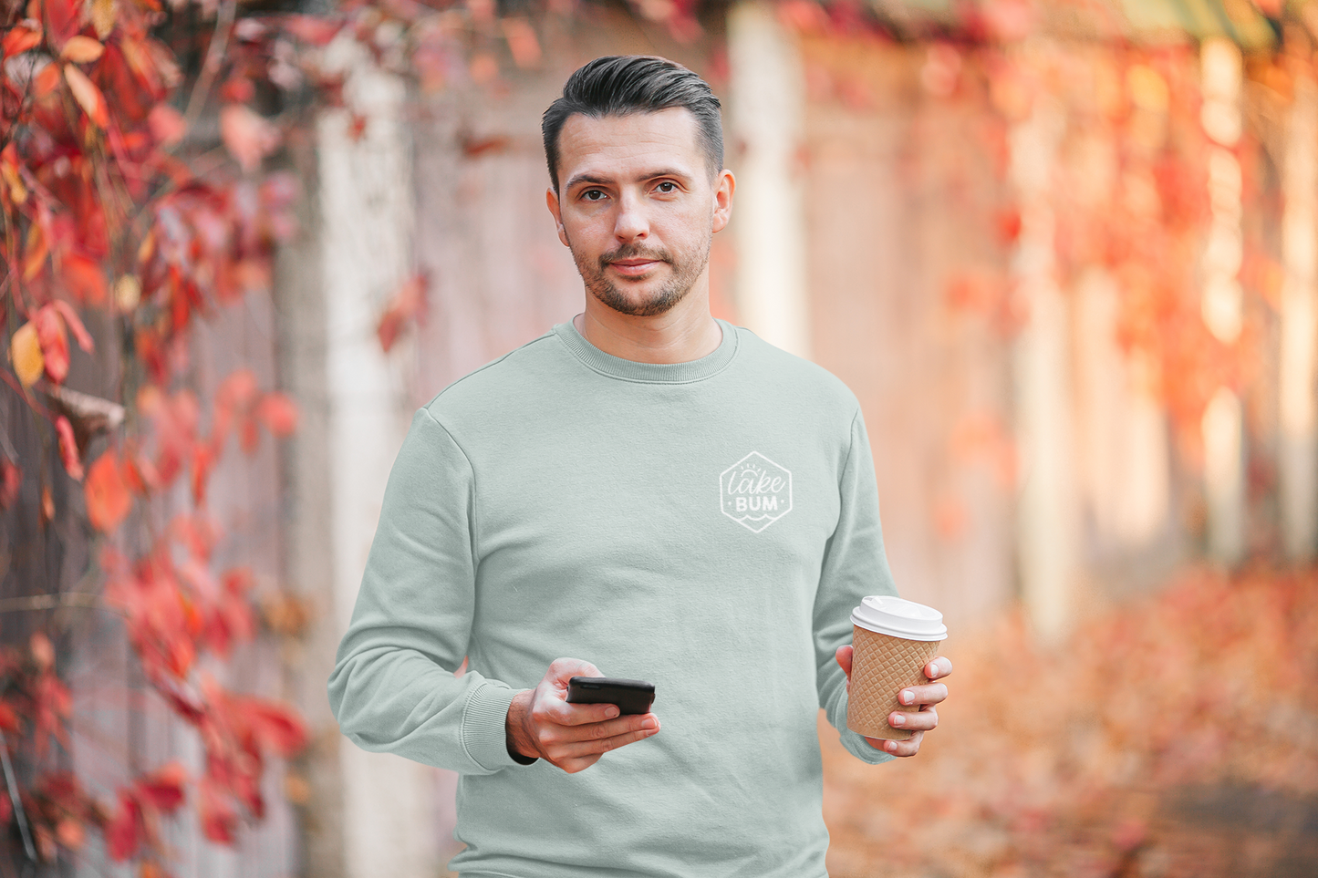 Life is Better at the Lake - Long Sleeve T-Shirt in heather dusty blue. The back of the shirt has a white design with the saying "Life is better at the lake" with lake waves, pine trees, and a loon all squared into a frame. The front has the saying "lake bum" with a lake wave and sun all boxed into a hexagon on the small square pocket (right). This is the front view of the shirt on a male model holding a phone and cup of coffee, surrounded by a fall leaf scenery.