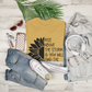 Rise Above The Storm Sunflower T-Shirt. The front has a half of a black sunflower on the left, with the right half having a quote of "rise above the storm and you will find the sunshine". This is a black design on a yellow shirt. This is the front view of the shirt folded on a wooden background stylized with other like items next to it.