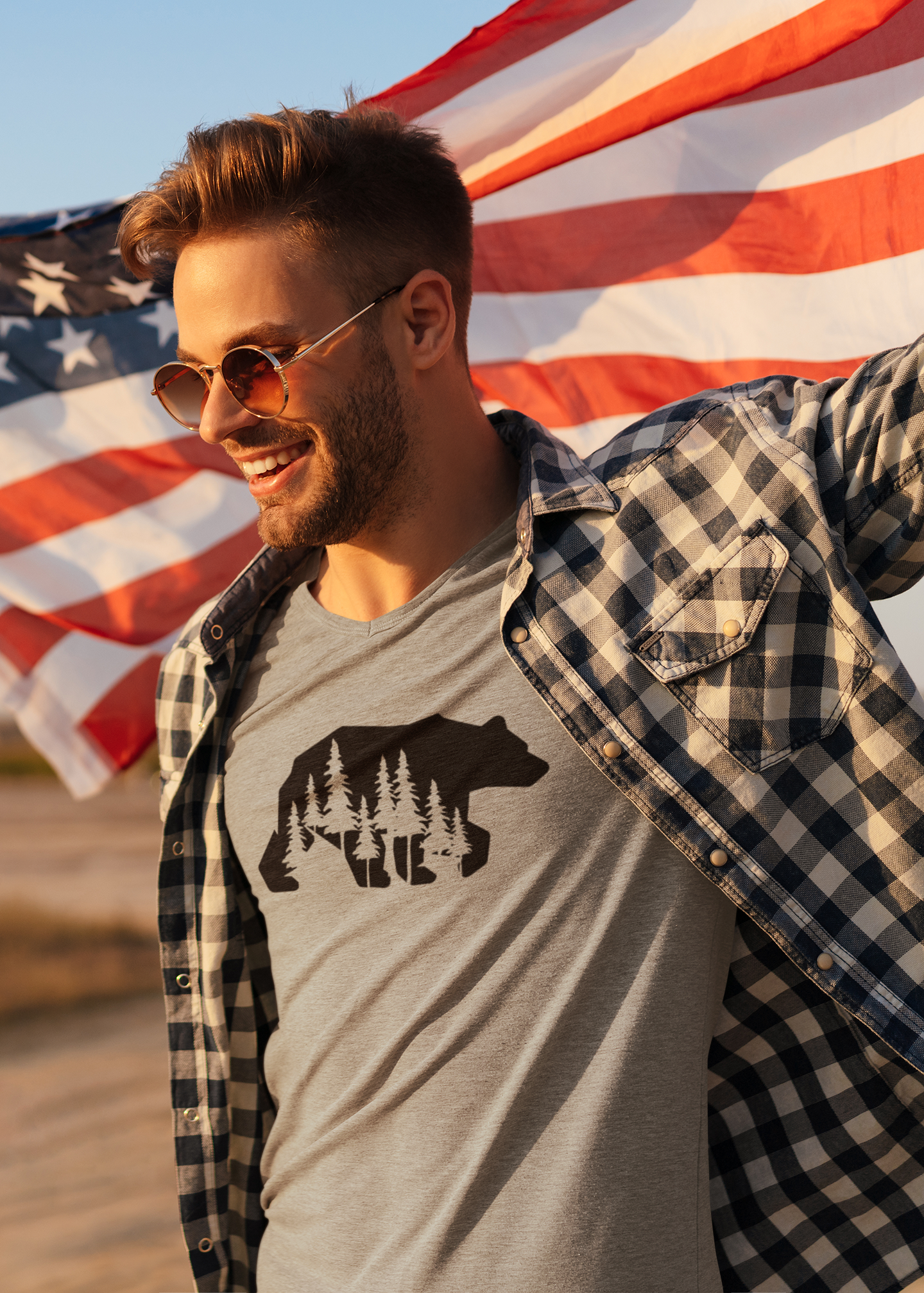 Woodsy Bear Silhouette - T-Shirt in gray with a black bear on it on a male model. The model has a black and white check plaid flannel, with sunglasses carrying an American flag behind his back.